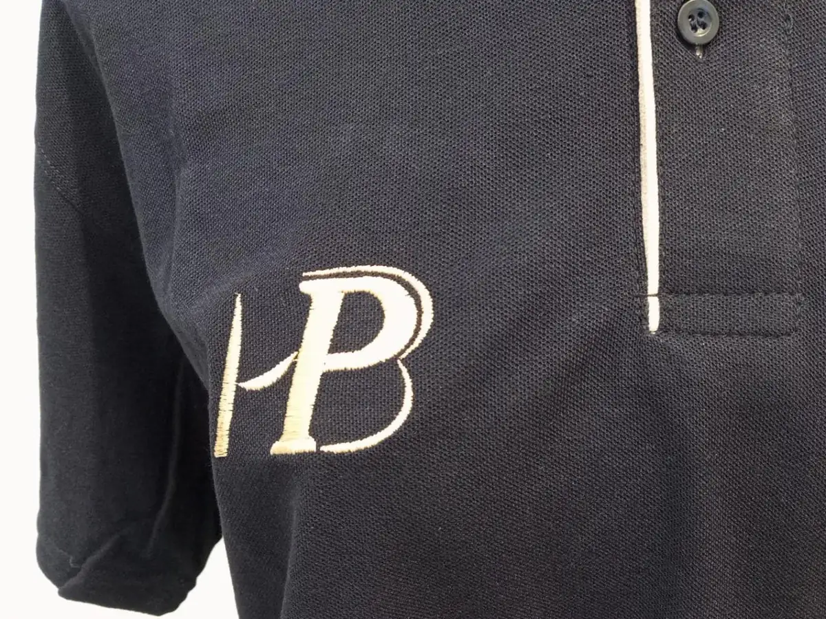 Iniciales bordadas en el pecho derecho de un polo negro, junto a la tapeta