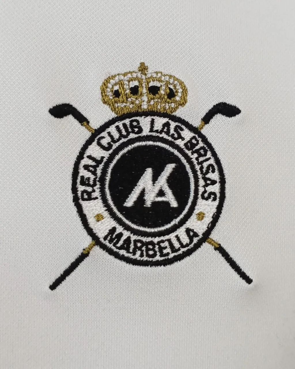 Detalle de bordado de escudo con hilo metalizado en el pecho de un polo blanco