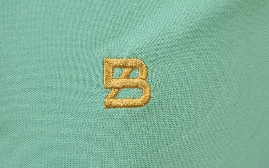 Detalle macro de bordado en relieve 3D con hilo dorado sobre polo verde